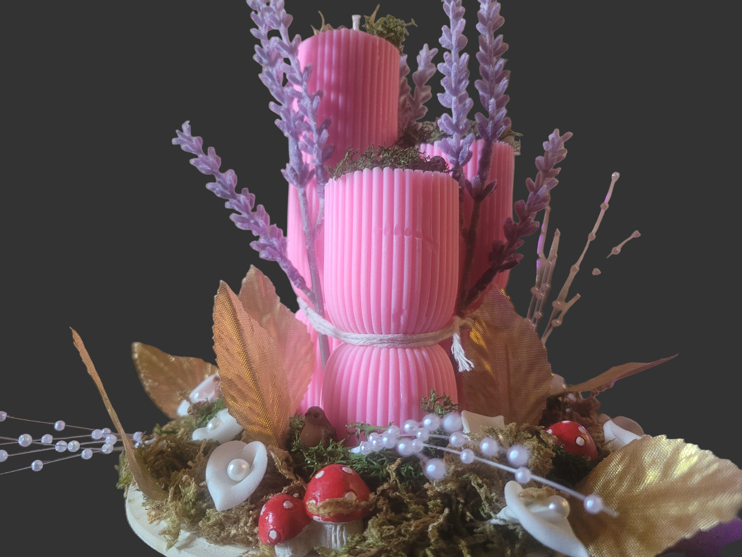 Decorative arrangement with pink ribbed candles, purple flowers, and greenery on a wooden surface.