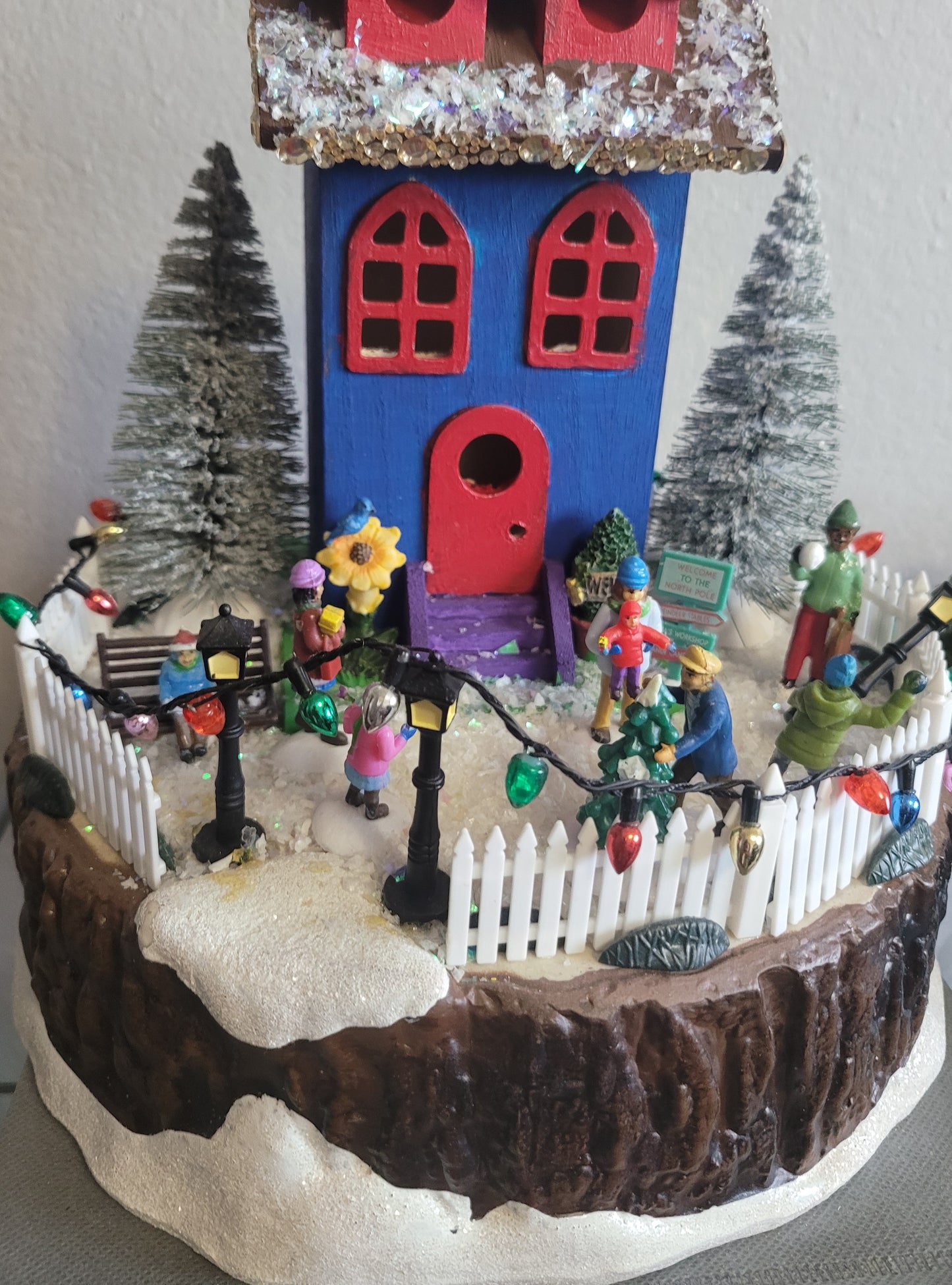 Decorative Christmas scene with a colorful house, trees, and small figures on a white background.