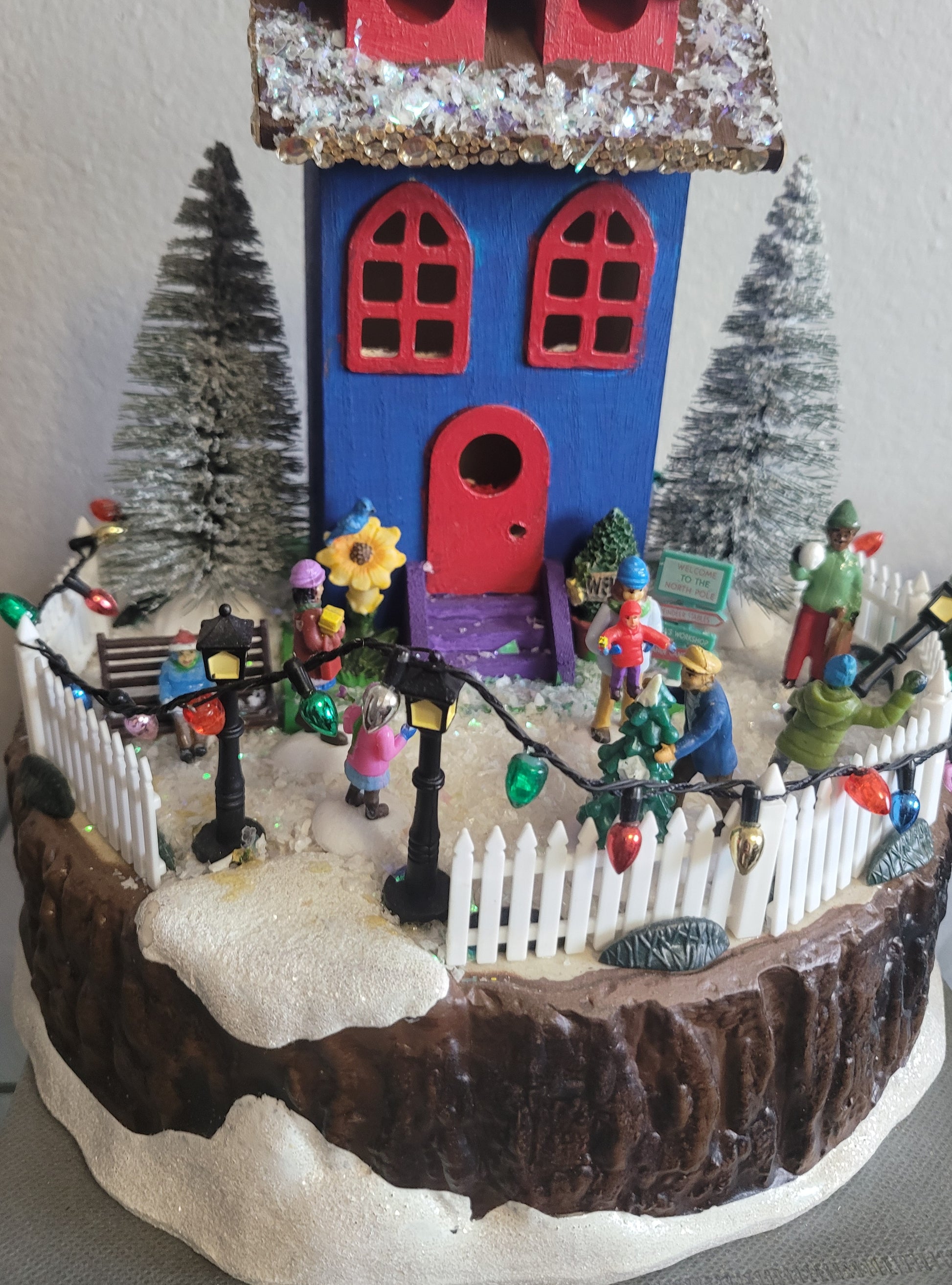 Decorative Christmas scene with a colorful house, trees, and small figures on a white background.