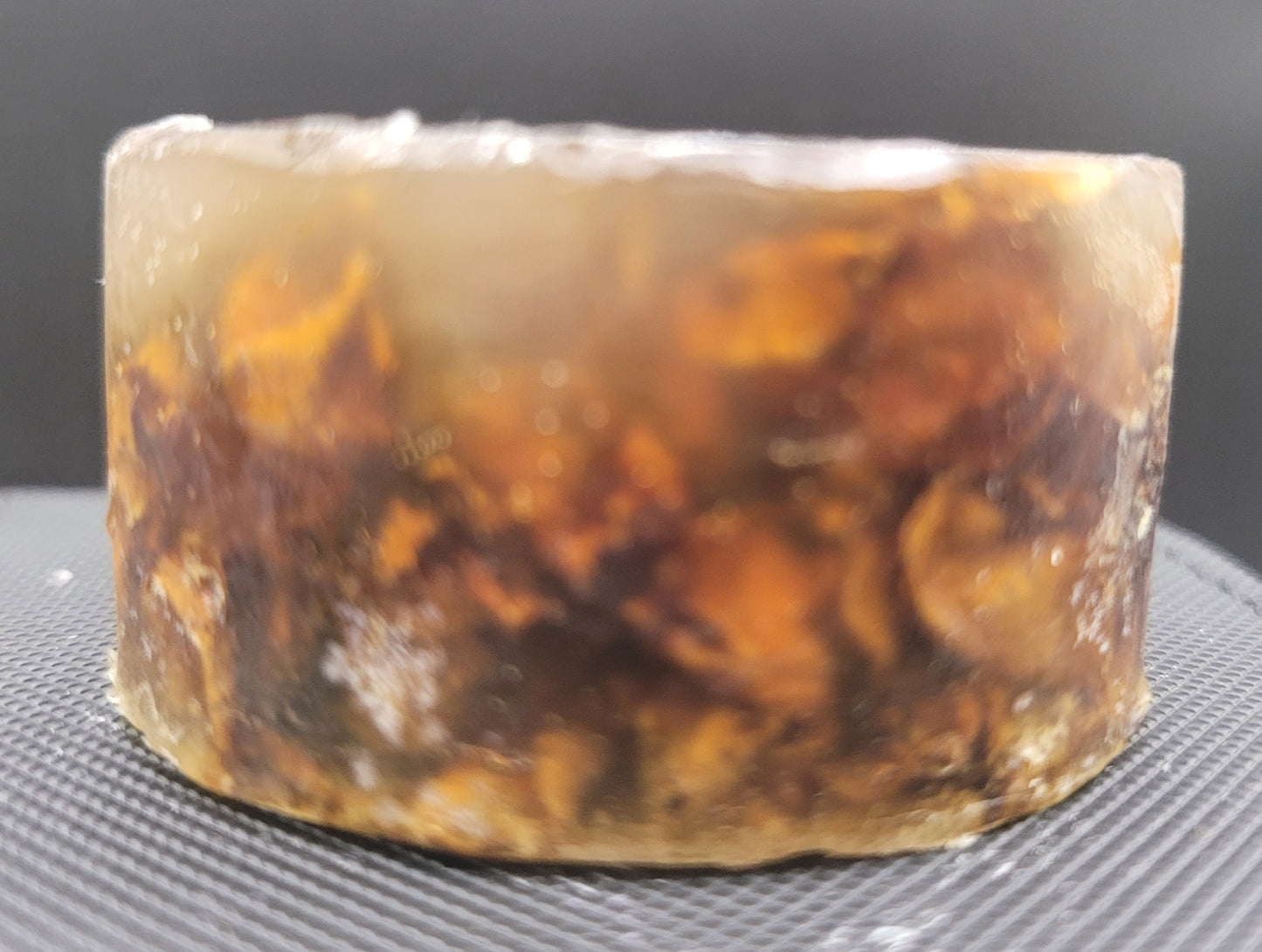 Cylindrical piece of amber colored soap with embedded rose petals on a dark surface